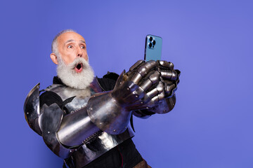 Armored elderly man in medieval knight costume taking a selfie in awe on a vibrant purple background