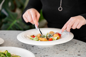 A close-up view of a person's hands holding a fork and knife, preparing to enjoy a healthy meal on a white plate. The dish features a colorful arrangement of salad and bite-sized portions.