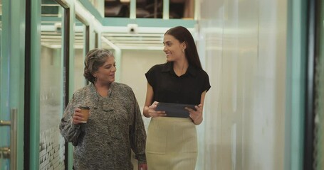 Indian older woman lady talk gossip young adult staff discuss work day time task indoor office place smiling female hold paper cup drink coffee expert girl hold tablet device walk at hall way space - Powered by Adobe