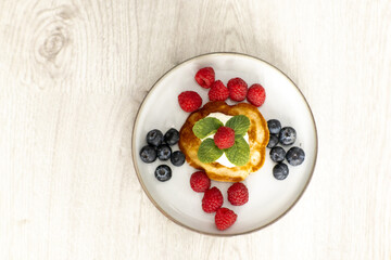 A stack of golden pancakes topped with fresh raspberries, blueberries, and mint leaves on a white plate. The delicious breakfast is presented on a light wooden table