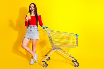 Stylish young woman with cheerful smile holding phone beside shopping cart against vibrant yellow...