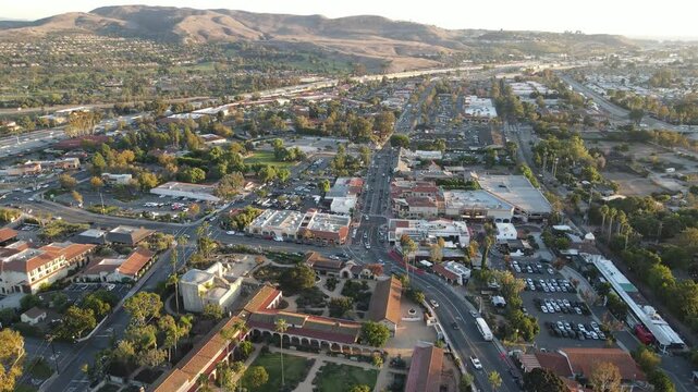 Aerial drone shot moving backward over San Juan Capistrano, CA, capturing historic architecture, bustling streets, and rolling hills. The Mission and surrounding buildings glow in golden hour light.