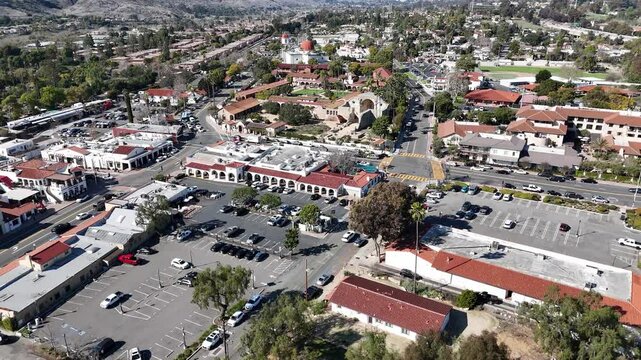 Aerial drone shot over San Juan Capistrano, CA, showcasing Mission San Juan Capistrano, historic Spanish-style architecture, and a vibrant downtown with shops, streets, and parking areas.
