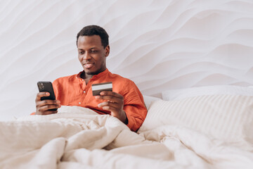 Happy black adult man lying comfortably in bed under a cozy blanket, using a smartphone and credit...