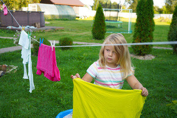 The backyard is peaceful as a young girl busily clips laundry, her golden hair catching the...