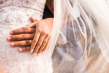 Noivo e noiva de mãos dadas com aliança de casamento e vestido na sessão pós wedding externa. 
