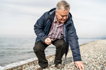Older male bending down to collect rocks on a pebble beach, engaging with nature. Represents retirement freedom, mental well-being, solitude, and connection to the earth.