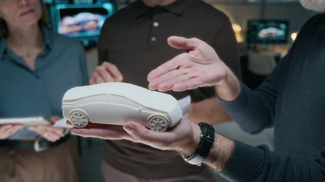 Medium closeup of hand of middle-aged male car engineer holding small-scale clay model of new crossover, pointing at exterior features while persuading colleagues during team meeting in office
