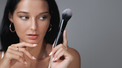 A beautiful woman inspects a makeup brush while highlighting her flawless skin and elegant...