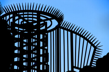 Steel silhouette of a metallic rotating turnstile and a fence with sharp spikes against the blue sky
