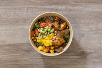 Overhead shot of quinoa bowl in an eco-friendly take-away container. Salmon, fresh mango cubes, edamame, cherry tomatoes, peanuts, mixed greens and quinoa. Placed on a wooden surface with copy-space