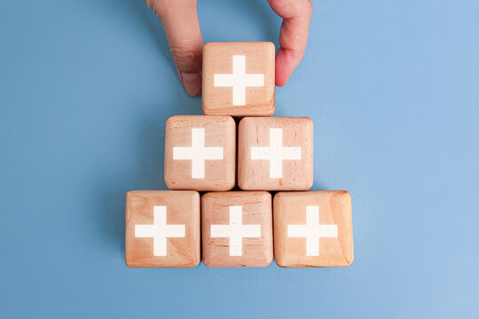 A hand places the top wooden block on a pyramid of six blocks, each marked with a white plus sign. The image symbolizes growth, teamwork, healthcare, and positive progress on a blue background.