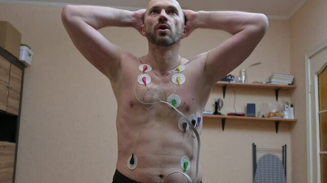 Man undergoing ECG/EKG cardiac monitoring at home with electrode patches on chest. Medical heart rhythm monitoring with electrocardiogram sensors. Holter monitor testing for heart health assessment.