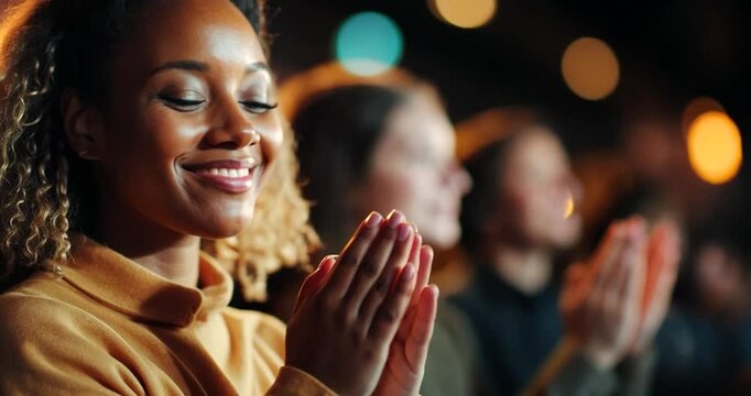 Close-up of a young woman with curly hair wearing a yellow sweater, smiling and clapping her hands in applause during a performance.  Warm lighting, blurred background of other audience members also