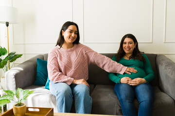Smiling doula gently touching the belly of an expecting mother, both seated comfortably on a sofa...