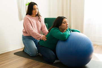 Young doula providing a soothing massage to a pregnant woman resting on a fitness ball during a supportive home visit