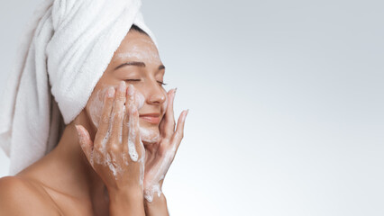 A serene woman with a towel wrapped around her head gently cleanses her face with foaming cleanser,...