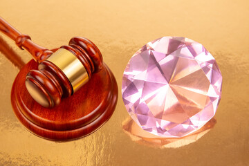 A wooden gavel with a gold band sits beside a large, sparkling pink diamond on a reflective gold...