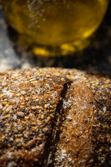 Vertical close up image of artisan whole grain rye bread next to a glass jar of oil on a dark wooden background with scattered flour.Nutritious food with a high fiber content. dark food