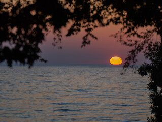 Sunset over Lake Ontario framed by the leaves of a tree sun dipping down over the horizon with a...