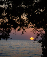 Fototapeta premium Sunset over Lake Ontario framed by the leaves of a tree sun dipping down over the horizon with a purple and orange sky shot in Grafton Ontario Canada backdrop or background for travel tourism Canada