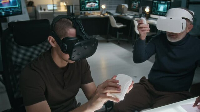 Medium shot of two male car designers sitting at desk in studio, using virtual reality headsets, viewing clay model, discussing improvements to new sedan modification and exterior shape