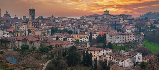 The old town of Bergamo, Italy, features Torre del Gombito, the Basilica of Santa Maria Maggiore, terracotta rooftops, and rolling hills at sunset.