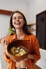 Happy young woman in an orange shirt enjoying a healthy salad in a bright kitchen, showcasing...