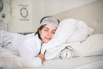 Young woman lying in bed wearing a sleep mask, with an alarm clock beside her, reflecting the...