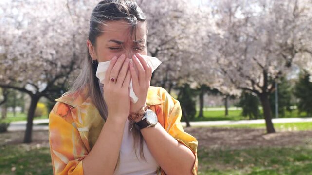 A woman in the park near the tree in spring blossom sneezing and blowing her nose in a paper tissue from a pollen allergy