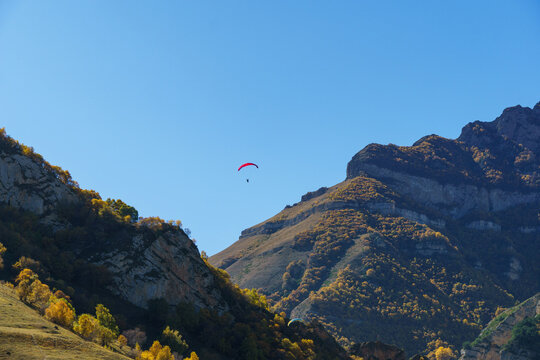 Paraglider floats serenely above lush, autumnal forest, surrounded by rugged cliffs and vibrant fall foliage. Fly Chegem paragliding site, El-Tyubu, Russia - October 7, 2024