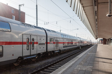Fototapeta premium A sleek silver double-decker passenger train with a red stripe is stationed at a quiet urban railway platform.