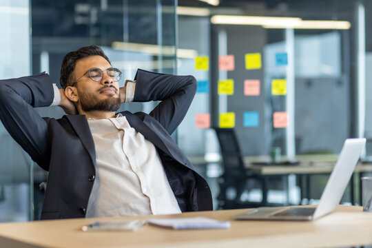 Businessman relaxing and resting inside the office. Man in business suit with hands behind head with closed eyes thinking and dreaming. Boss at workplace, working with laptop.