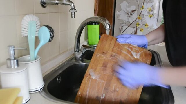 Woman is washing a wooden cutting board in a sink. She is wearing gloves and a blue cloth