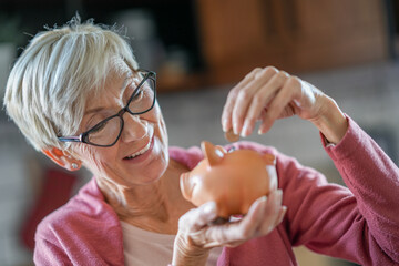 Senior woman smiling while placing a coin into a piggy bank, symbolizing financial savings and retirement planning. She enjoys financial security and careful money management in a cozy home setting.