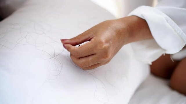 closeup of a woman&rsquo;s hand cleaning fallen hair from the bed and pillow caused by stress, hormonal changes, or health problems, dealing with hair loss at home