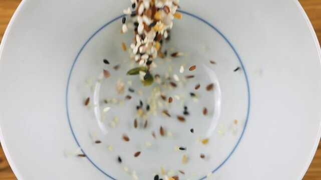 Salad seed mix is ​​poured into a white ceramic bowl. Sesame, flax, caraway, sunflower and pumpkin seeds. Top view. Slow motion.