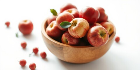 Obraz premium A wooden bowl brimming with ripe, red apples, complemented by scattered smaller red fruits on a pristine white background.