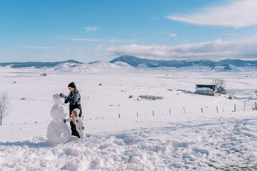 Little girl helps her mother build a snowman in a mountain valley. Side view © VolodymyrNadtochii
