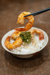 Chopsticks lifting a shrimp above a bowl of stir-fried shrimp and white rice.