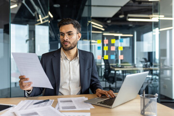 A thoughtful businessman reviews documents and uses his laptop, working in a modern office setting...