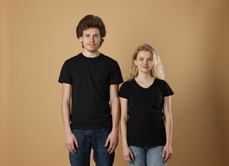 Young woman and man wearing blank black t-shirts on light brown background. Mockup for design