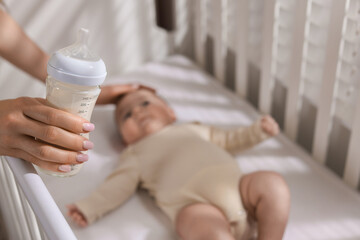 Mother with feeding bottle of milk near her little baby indoors, selective focus