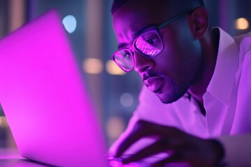 A man wearing glasses intensely focuses on his laptop screen in a dimly lit room.
