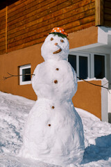 Snowman with a saucepan with red polka dots on his head stands near a wooden house