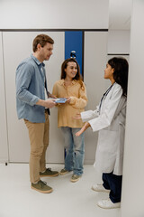 Fototapeta premium Female doctor standing and consulting a pregnant woman and a man standing next to her and smiling, in a hospital, during pregnancy