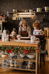Family, granddaughter standing at the table holding cookie cutters while grandmother stands next to her holding a rolling pin, grandson kneeling on a chair next to them, in the kitchen