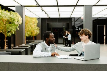 A male intern sits and puts his hand on the shoulder of a male employee who sits next to him and...