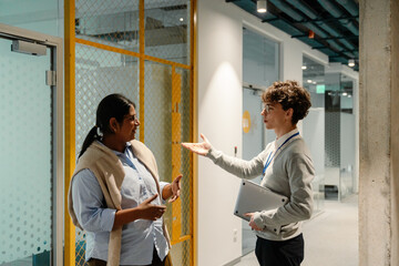 Male intern standing and holding a laptop while talking to a female employee standing next to him,...