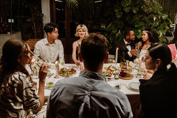 An adult Black groom with short dark hair drinks champagne with a young Black bride in a white dress at their wedding, while a group of diverse guests sits around a table with food in a restaurant.